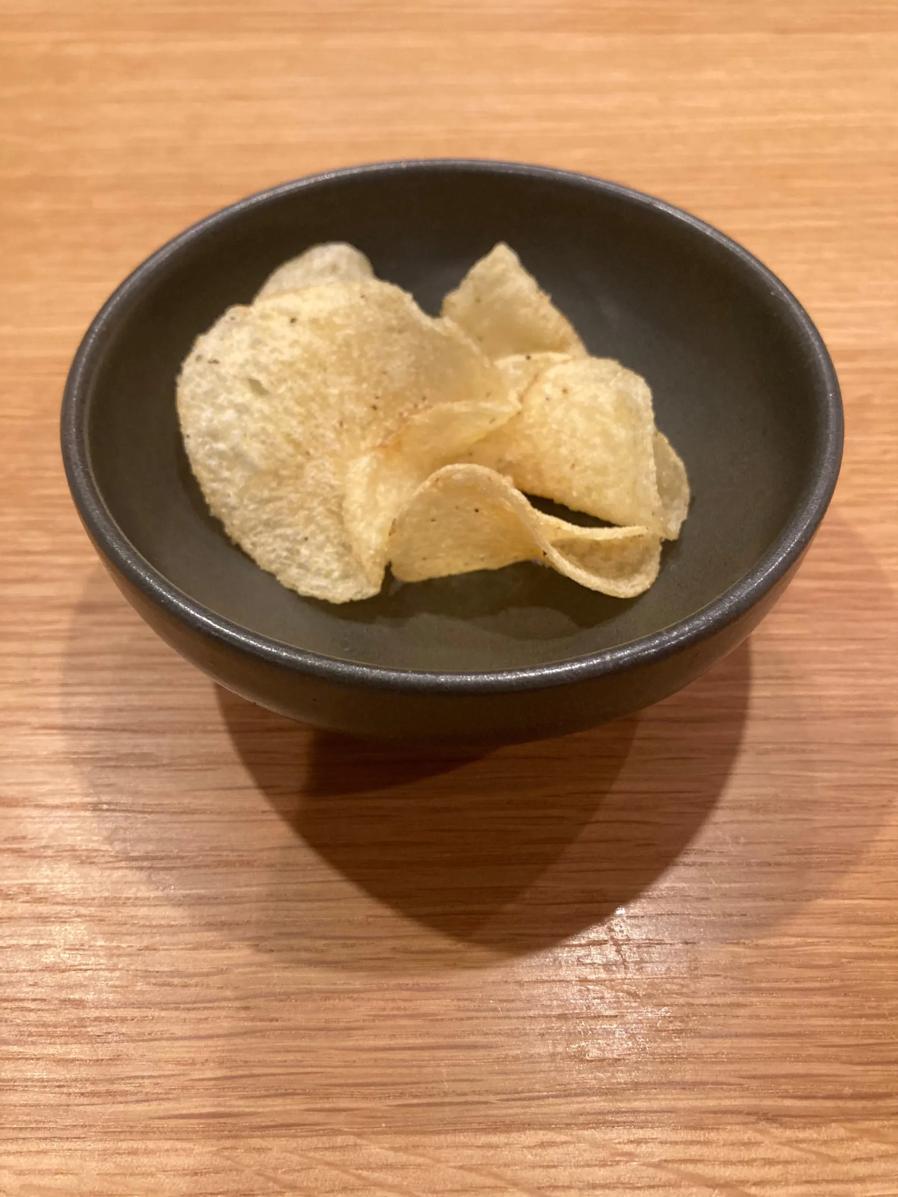 黒い器に盛った堅あげポテト ねぎ塩だれ味。厚みのある波打った形で、表面に黒こしょうの粒が見える